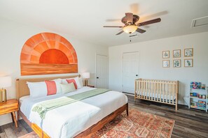 Ein Schlafzimmer mit einem Bett, einem Babybett, einem Deckenventilator, einer Tür und gerahmten Pflanzenbildern an der Wand.