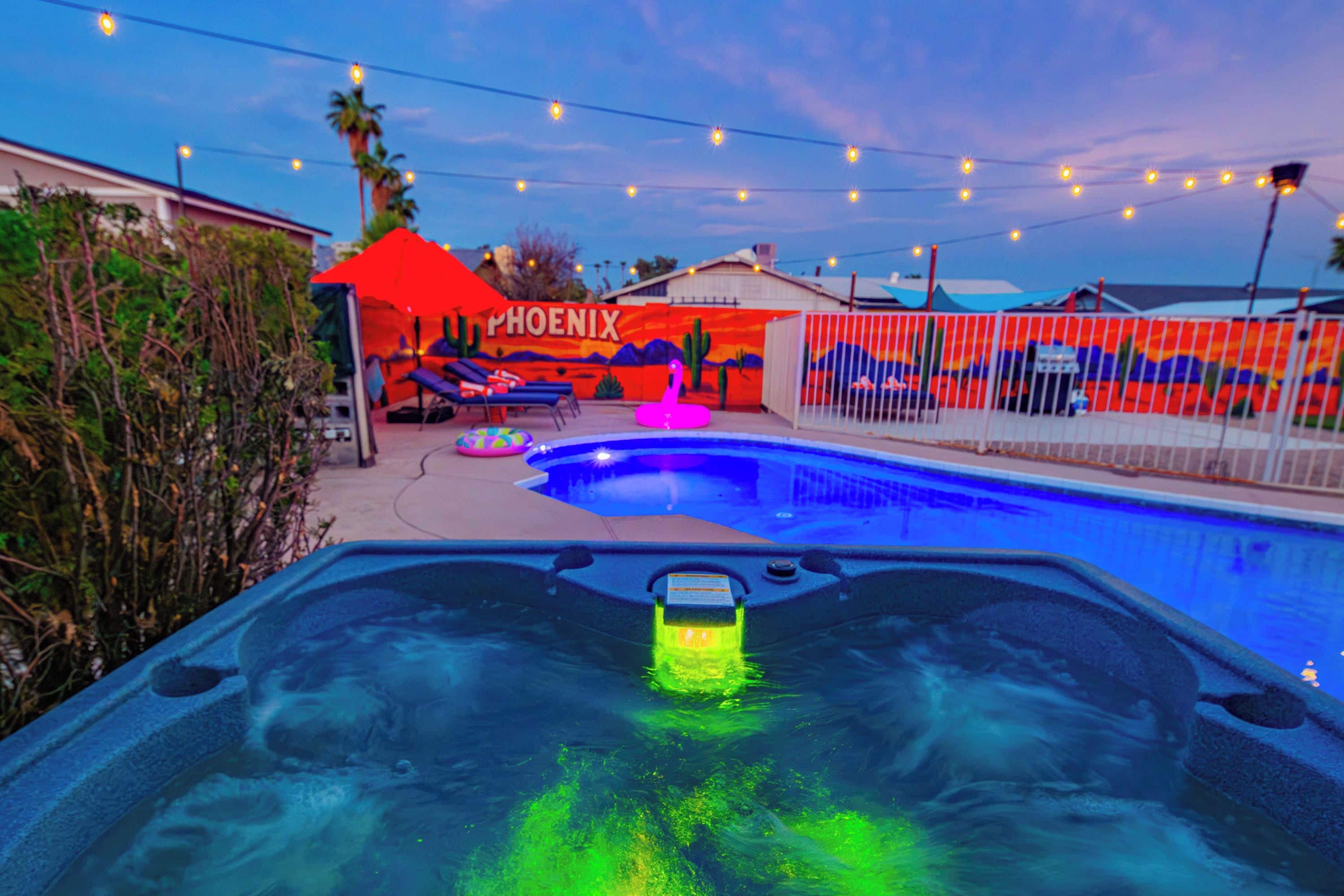 A swimming pool with a hot tub in the foreground, surrounded by a fence and a mural with the word 'PHOENIX'.