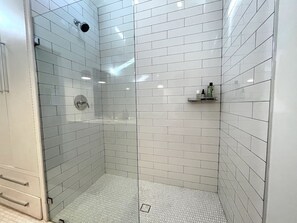 Et moderne badeværelse med hvide, rektangulære fliser, et glasbadeværelse og en lille hylde med toiletartikler.