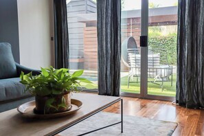 Una habitació amb un sofà, una taula de cafè amb una planta, i una porta corredissa de vidre que dona a una zona exterior amb taula i cadires.