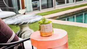Mesa redonda con una planta en maceta y un vaso con líquido anaranjado, posiblemente un cóctel, en un entorno junto a la piscina.