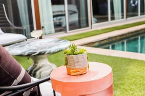 Et rundt bord med en potteplante og et glas appelsin-farvet drikkelse, sandsynligvis en cocktail, i en pool-omgivelse.
