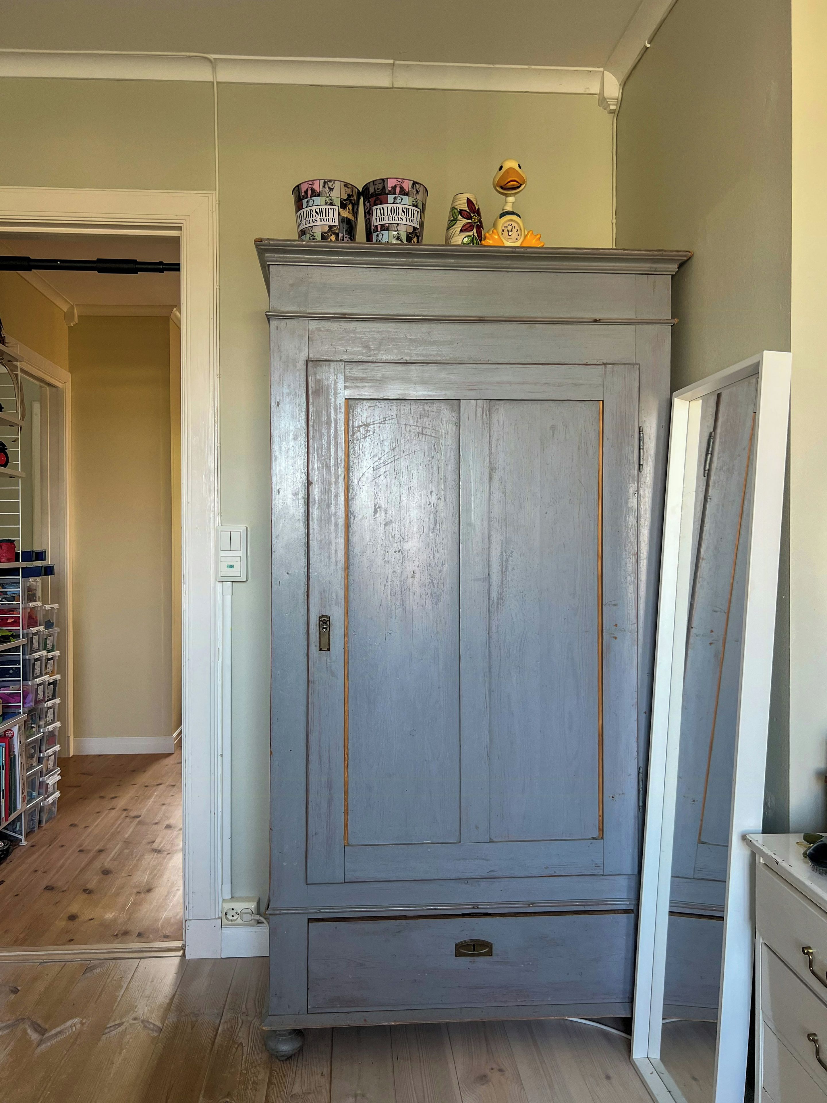A room with a large grey wardrobe, a wooden floor, and a shelf with decorative items.