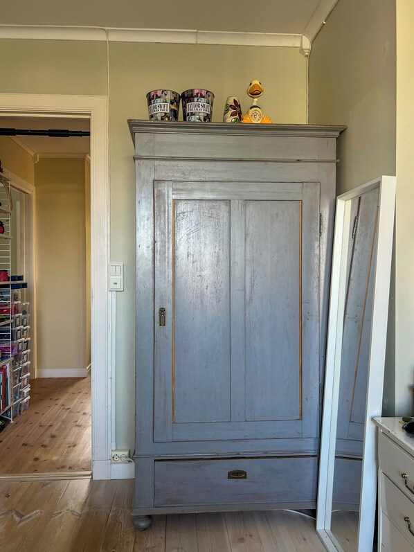 A room with a large grey wardrobe, a wooden floor, and a shelf with decorative items.