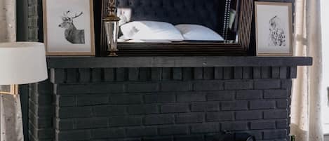A room with a black brick fireplace, a bed with a tufted headboard, and framed pictures on the mantel.