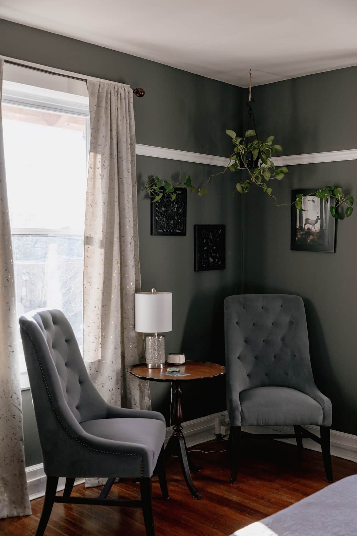A room with two upholstered chairs, a small round table with a lamp, and a window with curtains.