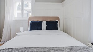 Quarto com cama, paredes brancas, janela com cortinas brancas e cabeceira branca.