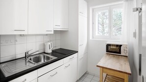 Cozinha moderna com armários brancos, bancada preta, pia de aço inoxidável, chaleira, micro-ondas e janela com vista para árvores.