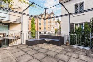 Een balkon met een zithoek, een bloempot en uitzicht op omliggende gebouwen.