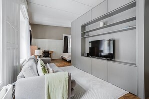 Uma sala de estar moderna com televisão de tela plana presa a uma parede cinza, um sofá com manta, uma mesinha com abajur e uma cama ao fundo.