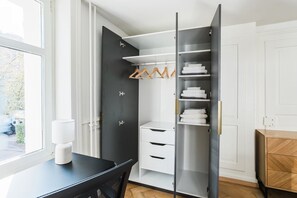 Ein Zimmer mit einer schwarzen Tür, einem weißen Schreibtisch mit Lampe, einem Holzschrank und einem Kleiderschrank mit Kleidung.
