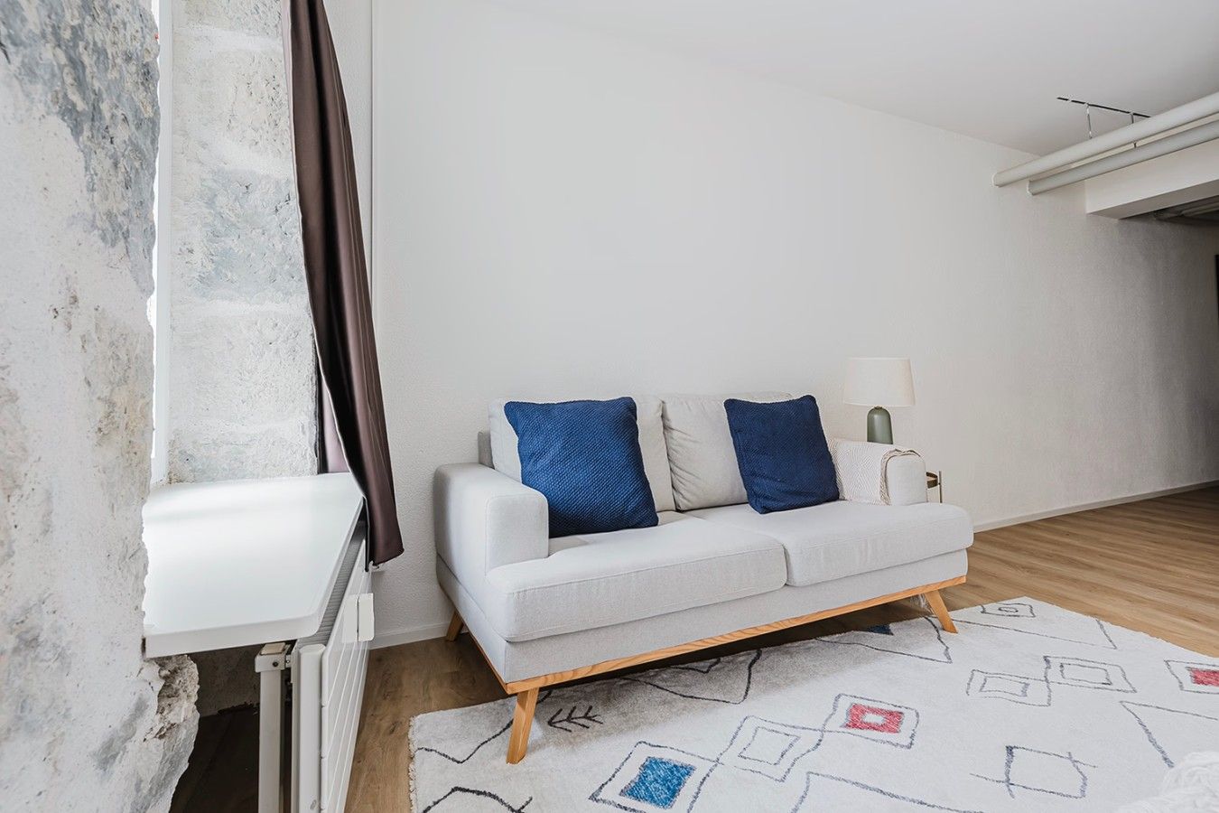 A modern living room with a white sofa, two blue cushions, a white side table, a lamp, and a patterned rug.