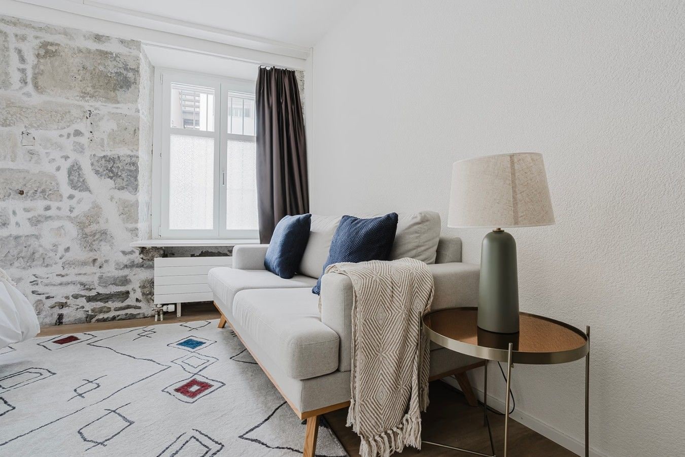 Una sala de estar moderna con pared de piedra, un sofá con cojines, un tapete estampado y una lámpara sobre un pedestal.