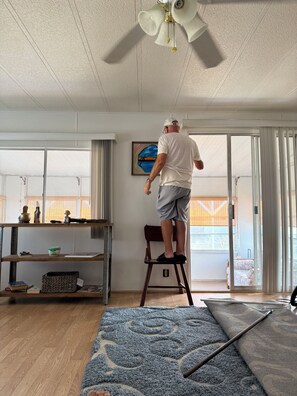 Una habitación con un ventilador de techo, una silla de madera, un tapete azul con estampado y un cuadro en la pared.