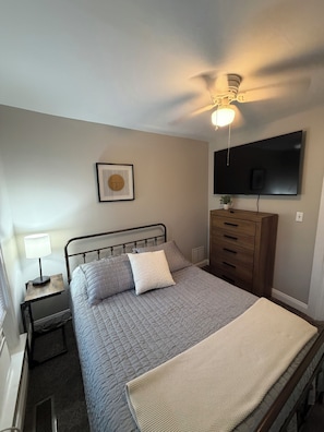 Een slaapkamer met een bed, nachtkastje met lamp, dressoir, plafondventilator, wandtelevisie en een lijst aan de muur.