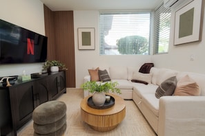 Ein modernes Wohnzimmer mit einem Flachbildfernseher, einer Couch, einem Couchtisch und einem großen Fenster mit Jalousien.