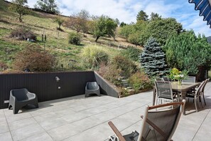 An outdoor patio area with a table and chairs, a dark fence, and a hilly landscape in the background.