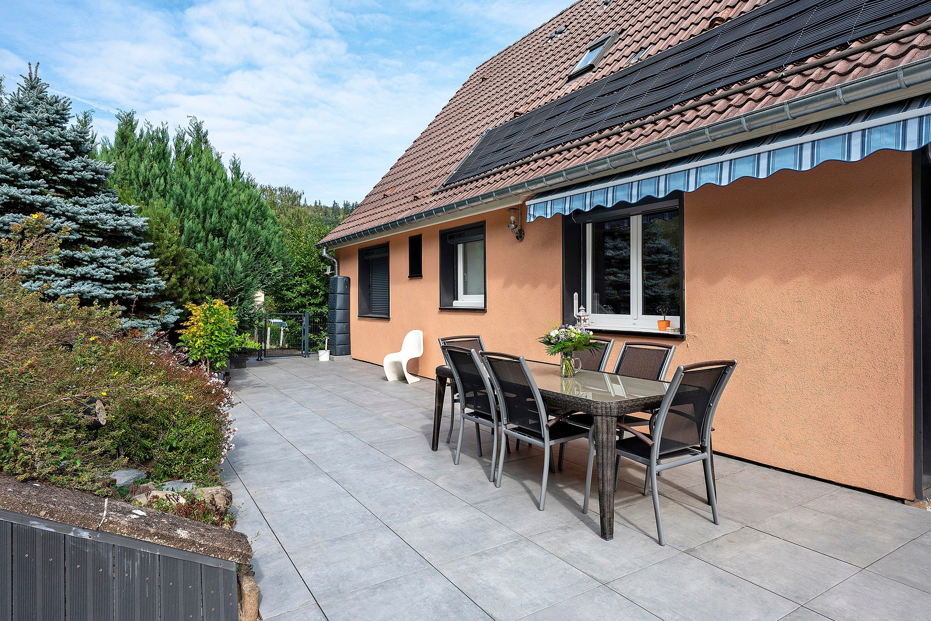 Une maison avec un toit en tuiles, une terrasse avec une table et des chaises, et un jardin avec des arbres et des arbustes.