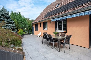 Una casa con techo de tejas, un patio con mesa y sillas, y un jardín con árboles y arbustos.