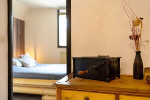 Um aparador de madeira com um rádio antigo e um vaso com plantas secas. Uma cama com roupa de cama branca é visível ao fundo.
