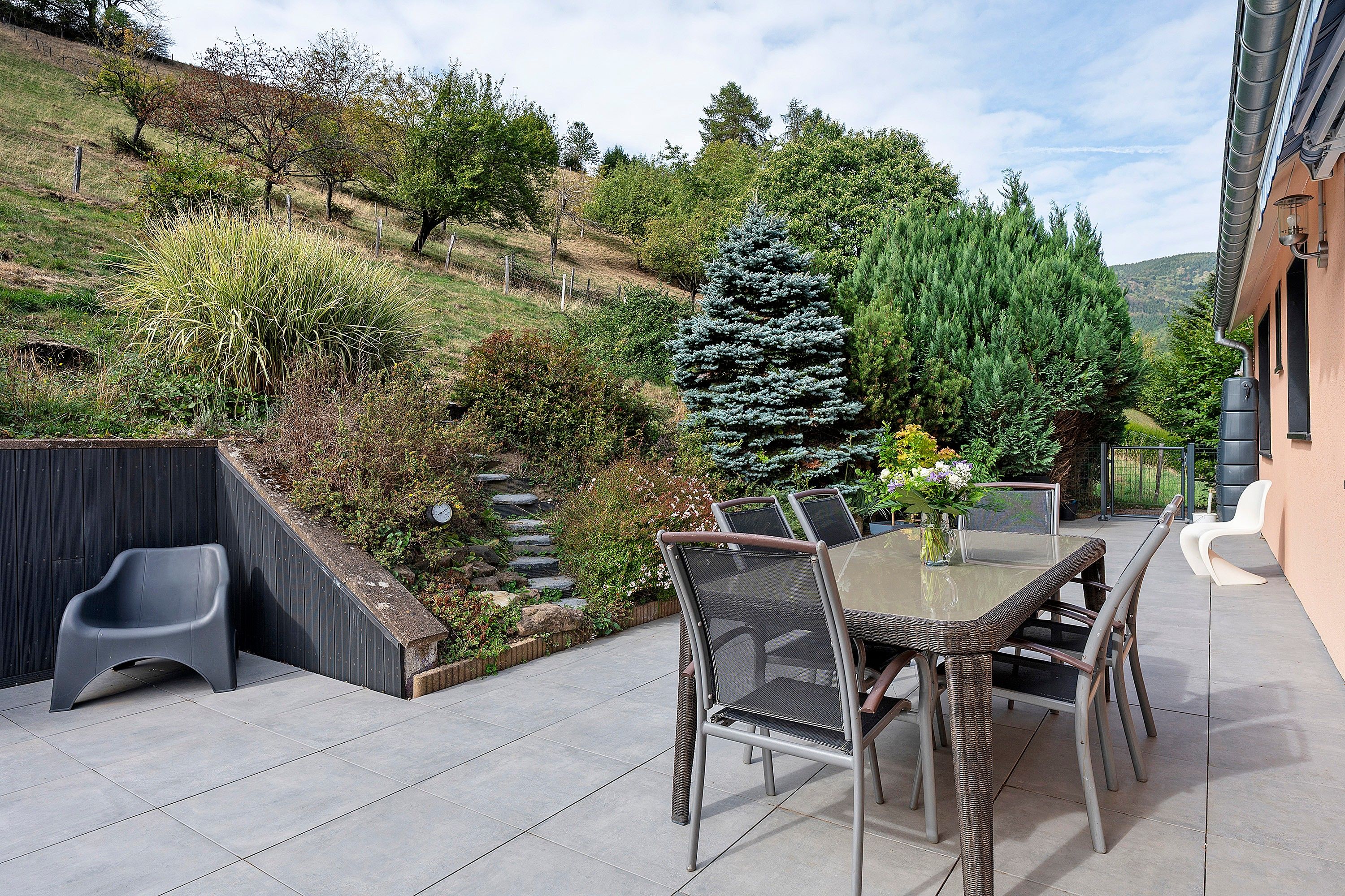 Um pátio externo com mesa e cadeiras, rodeado por um jardim ajardinado com diversas plantas e uma grande árvore conífera.
