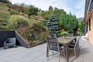 An outdoor patio with a dining table and chairs, surrounded by a landscaped garden with various plants and a large coniferous tree.