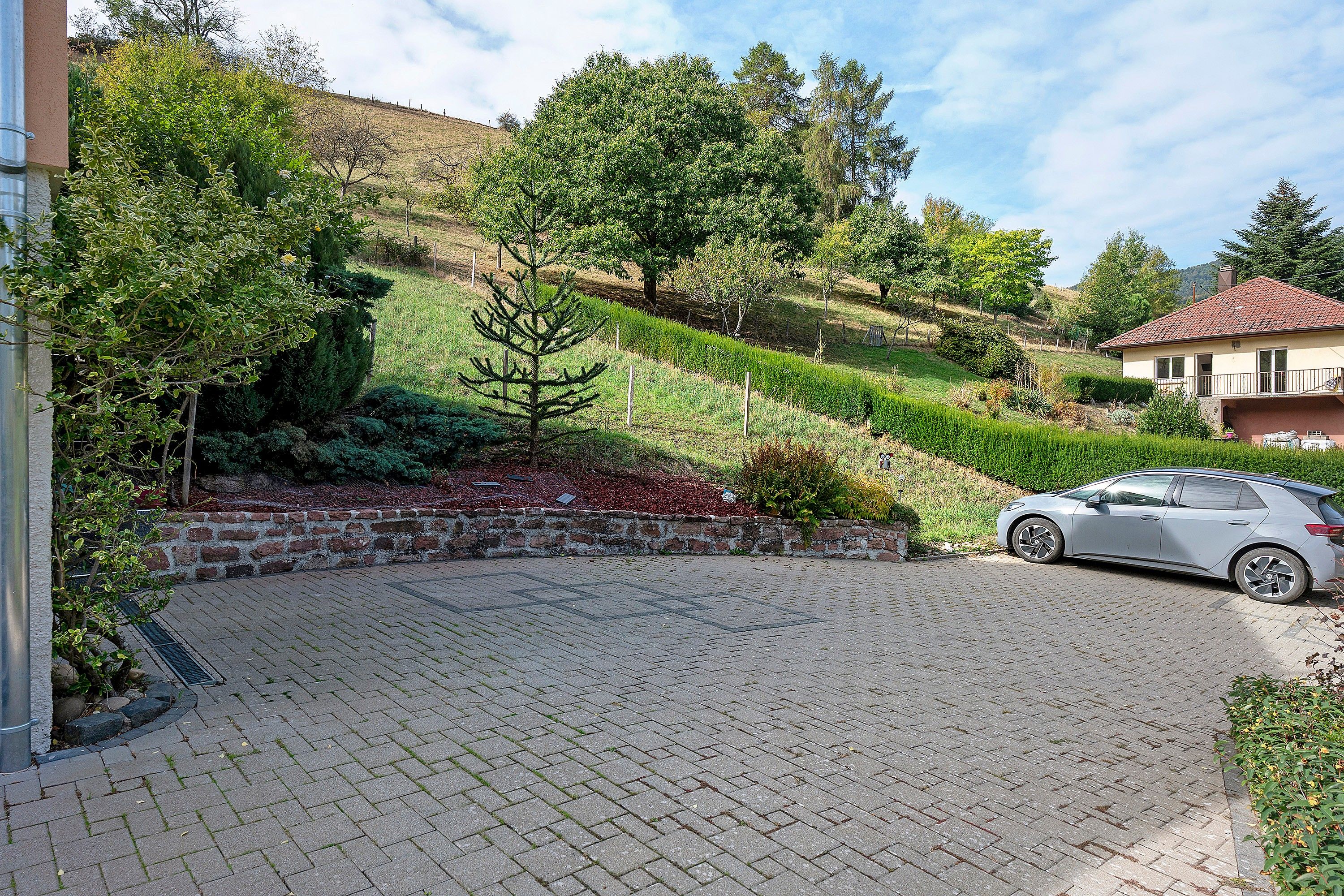 A paved driveway leading to a parked car, surrounded by landscaped gardens and a hillside.