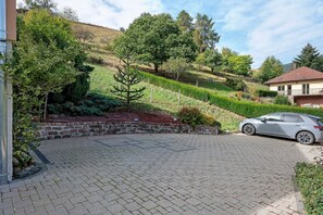 Um caminho de paralelepípedos leva a um carro estacionado, cercado por jardins bem cuidados e uma encosta.