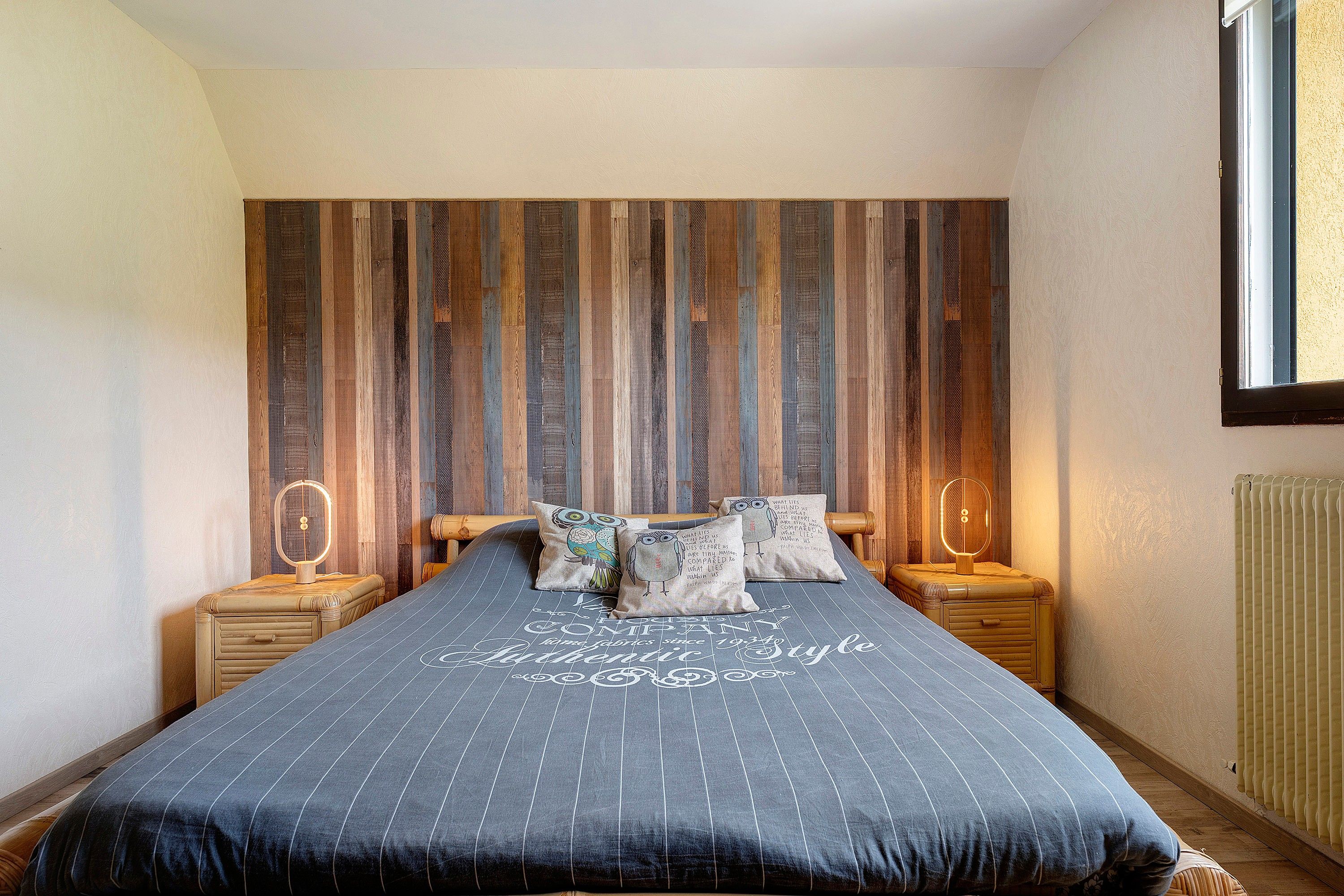 Une chambre à coucher comprenant un lit, deux tables de chevet, une fenêtre et un mur recouvert de lambris verticaux en bois.