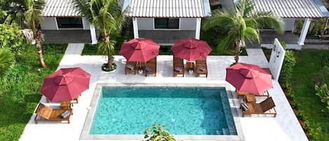 Una piscina rectangular con borde de azulejos, rodeada de sillones de madera y sombrillas rojas.
