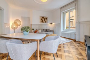 A modern living room with a dining table, grey upholstered chairs, a sofa, and a large window with a view. - Bright 3-bedroom apartment by the Limmat — tram at the door (Zürich)