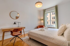 A bedroom with a bed, a desk, a chair, a round mirror, and a window with a view of buildings. - Bright 3-bedroom apartment by the Limmat — tram at the door (Zürich)