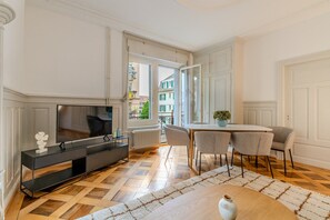 Ein modernes Wohnzimmer mit einem Flachbildfernseher, einem Esstisch mit vier Stühlen und einem großen Fenster mit Blick auf Gebäude.