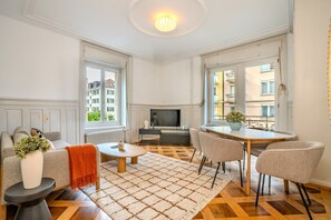A modern living room with a sofa, coffee table, and dining table. There is a TV and a window with a view of buildings. - Bright 3-bedroom apartment by the Limmat — tram at the door (Zürich)