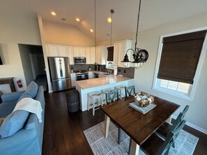 A modern kitchen with stainless steel appliances, a dining area with a wooden table and chairs, and a living room with a blue sofa. - Coastal Dream at Ortley Beach (Seaside Heights)