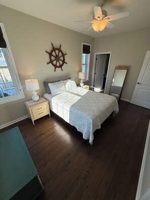 Une chambre à coucher comprenant un lit, une table de chevet, une lampe, un ventilateur de plafond, un élément de décoration représentant un gouvernail de bateau, une fenêtre et une porte.