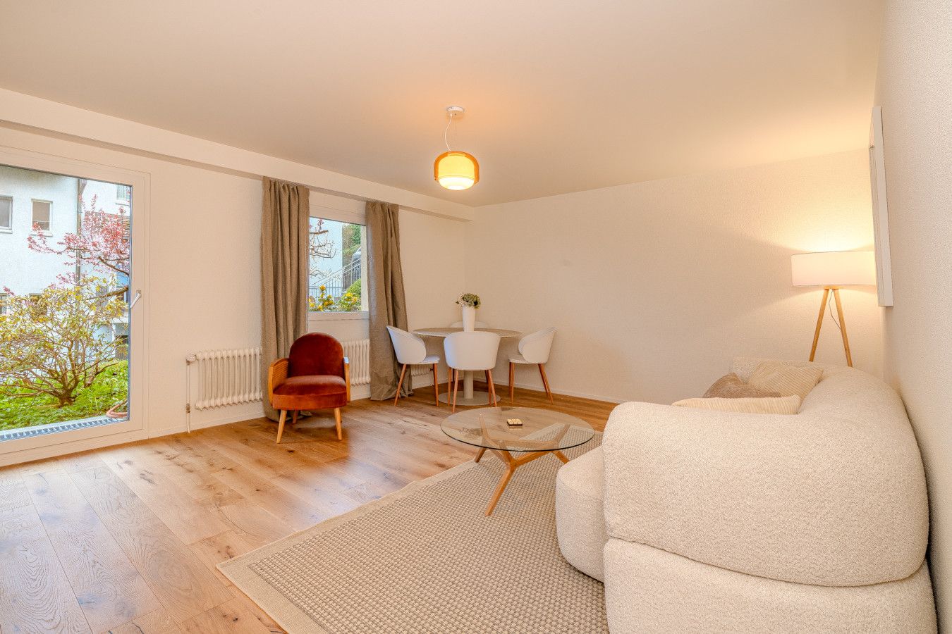 Ein modernes Wohnzimmer mit einer Couch, einem roten Sessel, einem Esstisch mit Stühlen, einer Lampe und Blick auf Grünfläche im Freien.