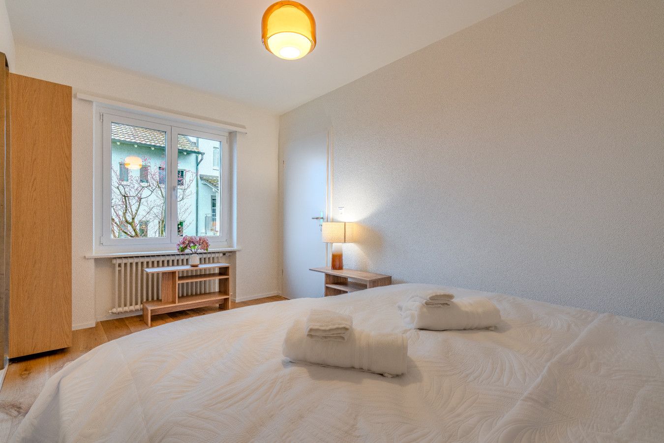 Ein Schlafzimmer mit einem großen Bett, einem Fenster mit Blick auf Gebäude, einem Nachttisch mit einer Lampe und einem Heizkörper.