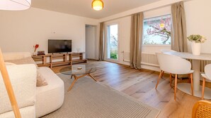 A modern living room with a flat-screen TV, a sofa, a glass coffee table, and a dining area with chairs. - Bright 1-BR with garden view & parking · Kilchberg (Kilchberg)