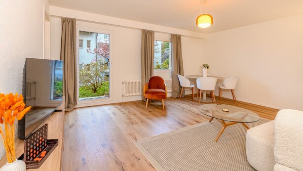 A modern living room with a television, a red chair, a dining table with chairs, and a large window with curtains. - Bright 1-BR with garden view & parking · Kilchberg (Kilchberg)