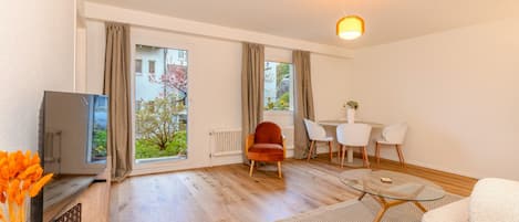 Ein modernes Wohnzimmer mit Fernseher, einem roten Sessel, einem Esstisch mit Stühlen und einem großen Fenster mit Vorhängen.