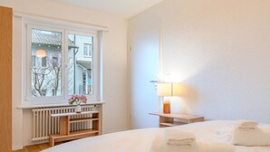 A hotel room with a large bed, a bedside table with a lamp, a window with a view of buildings, and a radiator. - Bright 1-BR with garden view & parking · Kilchberg (Kilchberg)