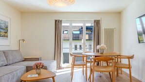 Una sala de estar moderna con sofá, mesa de centro, mesa de comedor y sillas. Hay un ventanal con cortinas, un radiador y una lámpara del techo.