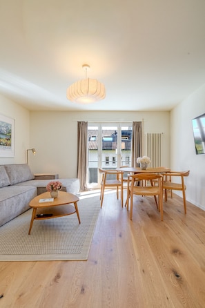 Ein modernes Wohnzimmer mit Sofa, Couchtisch, Esstisch und Stühlen. Ein großes Fenster mit Vorhängen, eine Heizung und eine Deckenleuchte sind ebenfalls vorhanden.
