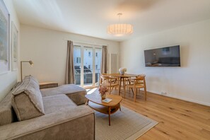 A modern living room with a sofa, dining table, chairs, a TV, and a large window with curtains. - Garden-level 2-BR with parking · Kilchberg (Kilchberg)