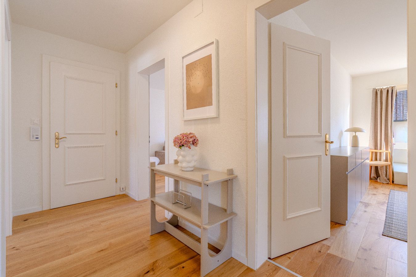 Ein Flur mit einer weißen Tür, einem weißen Regal mit Blumen in einer Vase und einem gerahmten Bild an der Wand.