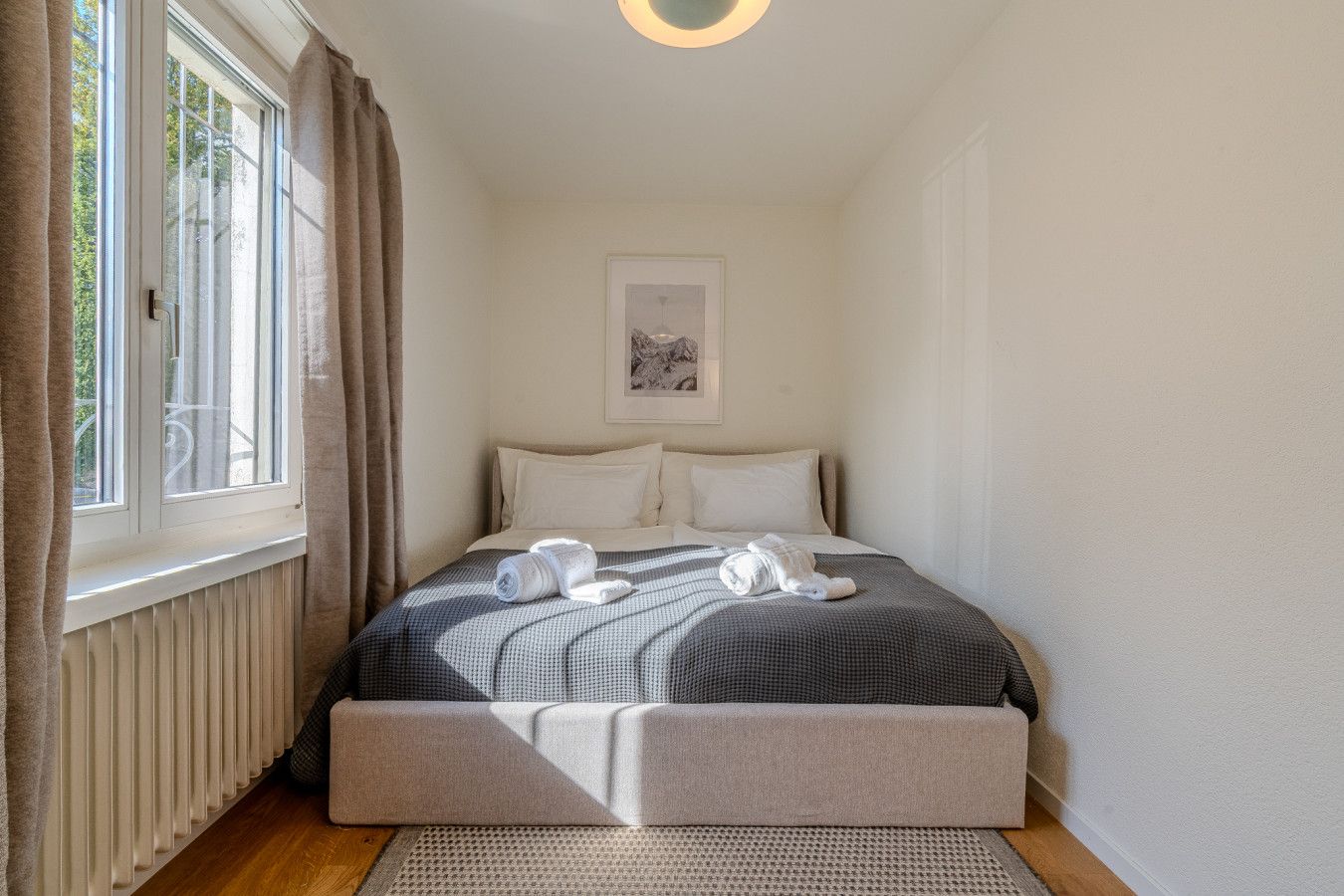 Ein Schlafzimmer mit einem Bett, einem Heizkörper, einem Fenster mit Vorhängen und einem gerahmten Bild an der Wand.