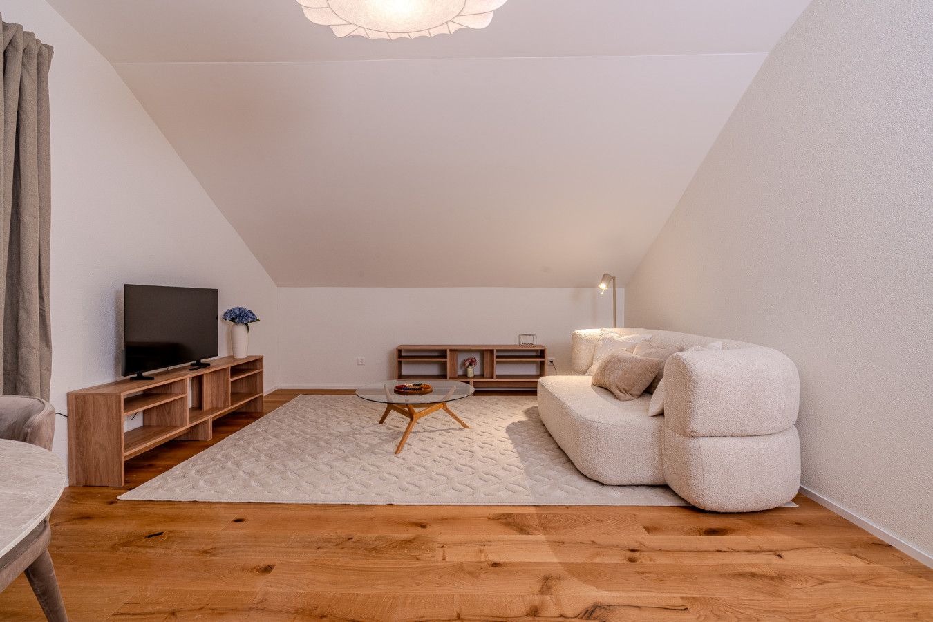 Ein modernes Schlafzimmer im Loft-Stil mit schrägverdeckter Decke, einem Flachbildfernseher, einer Holzeinrichtungsanlage, einem runden Glastisch, einem beigen Sofa und einem gemusterten Teppich.