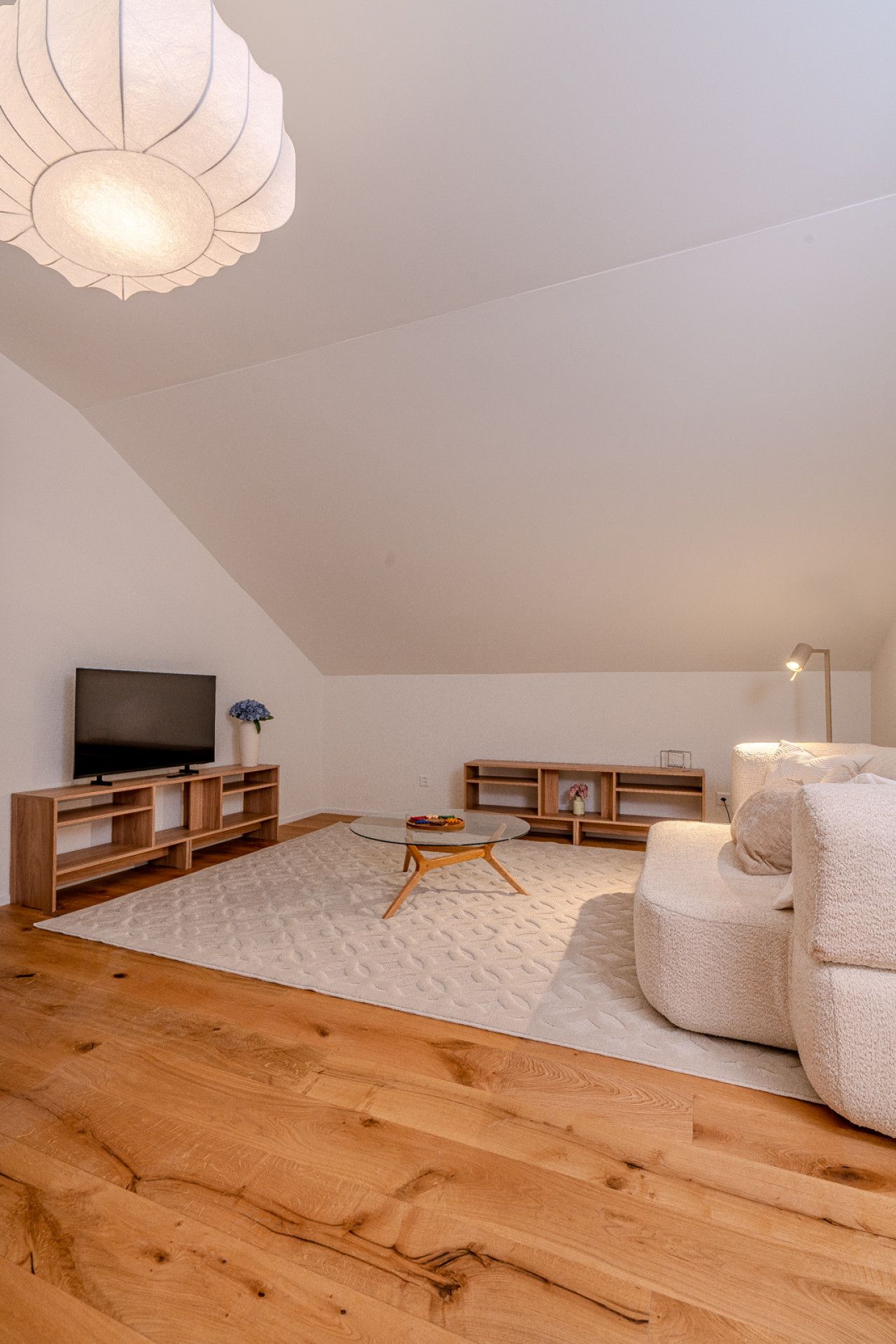 A modern living room with a flat-screen TV, wooden furniture, a round glass table, and a ceiling light fixture.