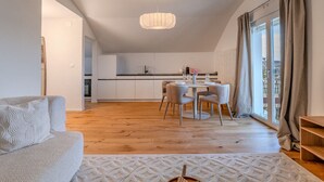 Ein modernes Wohnzimmer mit einer Couch, einem Esstisch mit Stühlen, einer Küche im Hintergrund und einem großen Fenster mit Vorhängen.